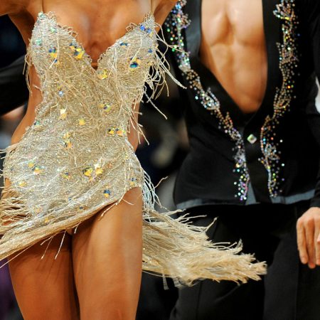 Tanzpaar auf Turnier der Lateinamerikanischen Tänze. Sie träg ein goldfarbenes, kurzes Kleid und steht vor ihrem Herren in schwarzer Kleidung. Im Hintergrund sind weitere Paare zu erkennen.